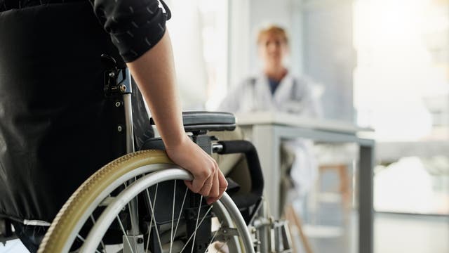Eine Person im Rollstuhl sitzt in einem hellen Raum und hält das Rad des Rollstuhls. Im Hintergrund sitzt eine unscharf dargestellte Person in einem weißen Kittel an einem Tisch, möglicherweise ein Arzt oder medizinisches Fachpersonal. Die Szene vermittelt eine medizinische oder pflegerische Umgebung.