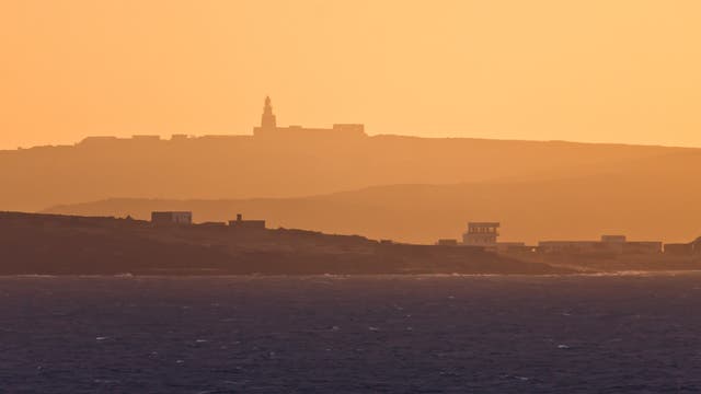 Eine Küstenlandschaft bei Sonnenuntergang. Im Vordergrund ist das Meer zu sehen, während im Hintergrund eine hügelige Landschaft mit mehreren Gebäuden, darunter ein Leuchtturm, in warmen Orangetönen beleuchtet wird. Die Szene vermittelt eine ruhige und friedliche Atmosphäre.