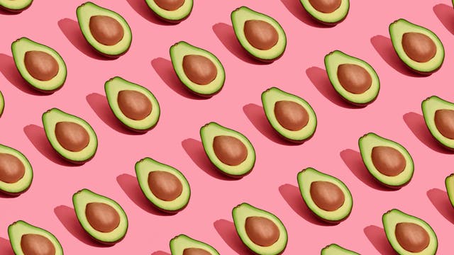 Mehrere Hälften von Avocados mit Kernen sind gleichmäßig auf einem rosa Hintergrund angeordnet. Die Avocados sind in diagonalen Reihen platziert, und jede Hälfte wirft einen Schatten. Die Komposition erzeugt ein modernes und frisches Muster.