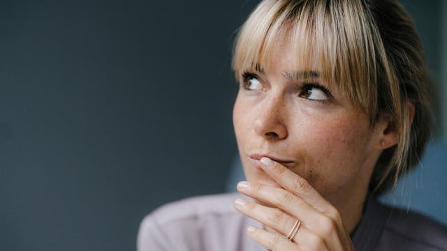 Eine Frau mit blonden Haaren schaut nachdenklich zur Seite. Sie trägt einen hellen Pullover und hat ihre Hand an ihr Kinn gelegt. Der Hintergrund ist dunkel und unscharf, was den Fokus auf ihr Gesicht lenkt.