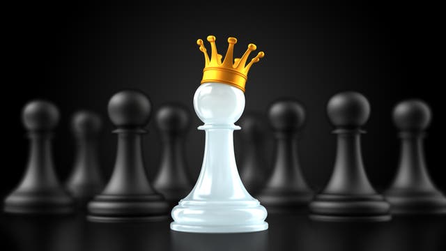 Ein weißer Schachbauer mit einer goldenen Krone steht im Vordergrund, umgeben von unscharfen schwarzen Bauern im Hintergrund. Die Krone symbolisiert Macht und Aufstieg, während der Kontrast zwischen dem weißen Bauern und den schwarzen Figuren die Themen Führung und Gefolgsleute hervorhebt.