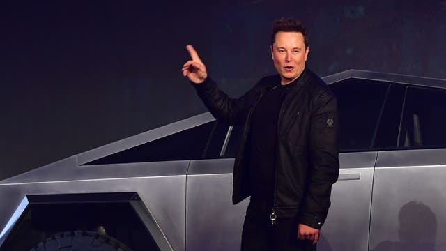 Tesla-CEO Elon Musk steht vor einem futuristisch aussehenden Fahrzeug mit kantigem Design und großen Reifen, einem Tesla Cybertruck. Er hebt einen Finger, als ob er etwas erklärt oder präsentiert. Das Fahrzeug hat eine silberne, metallische Oberfläche und leuchtende Scheinwerfer.