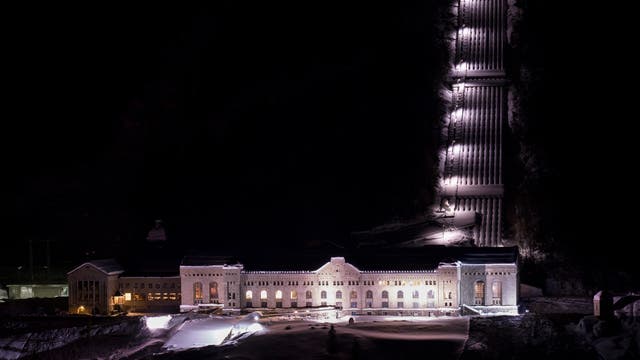 Ein großes, beleuchtetes Gebäude bei Nacht, umgeben von Schnee. Die Architektur des Gebäudes ist historisch und symmetrisch, mit zahlreichen Fenstern und einem zentralen Eingang. Rechts im Bild sind nebeneinander mehrere beleuchtete Rohre erkennbar, die von einem steilen Berg herunterführen. 