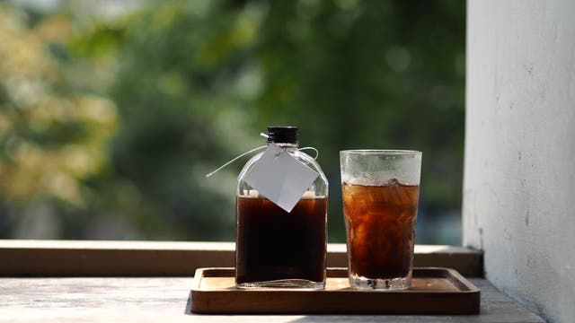 Eine Glasflasche mit dunklem Getränk steht neben einem Glas mit kaltem Kaffee und Eiswürfeln auf einem Holztablett in einer Fensteröffnung. Im Hintergrund sind unscharfe grüne Blätter zu sehen.