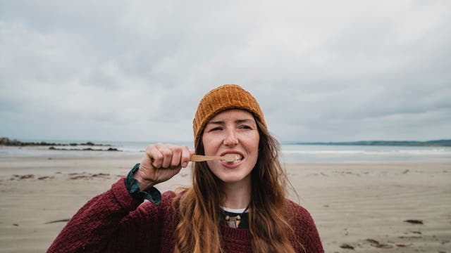 Eine Frau mit langen, rotbraunen Haaren und einer orangefarbenen Mütze putzt sich mit einer Bambuszahnbürste die Zähne. Sie trägt einen roten Pullover und steht an einem bewölkten Strand vor dem Meer. Im Hintergrund sind Wellen zu sehen.