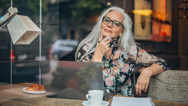 Eine Person sitzt in einem Café an einem Holztisch mit einem Laptop, einem Notizblock und einem Stift in der Hand. Auf dem Tisch stehen eine Tasse Kaffee und ein Croissant. Die Person schaut nachdenklich. Im Hintergrund sind verschwommene Straßenszenen zu sehen.