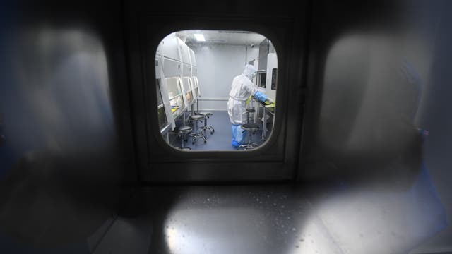 Eine Person in einem weißen Schutzanzug und blauen Überschuhen arbeitet in einem Labor. Sie steht an einem Labortisch und hantiert mit Geräten. Der Raum ist durch ein Fenster von außen sichtbar und enthält mehrere Arbeitsstationen mit Hockern. Die Umgebung wirkt steril und gut beleuchtet.