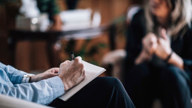 Eine Person sitzt mit einem Notizblock auf dem Schoß und schreibt mit einem Stift. Im Hintergrund ist eine weitere Person unscharf zu sehen, die möglicherweise in ein Gespräch vertieft ist. Die Szene deutet auf eine Beratung oder ein Interview hin. Der Raum wirkt gemütlich und professionell.
