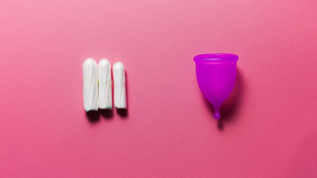 Drei weiße Tampons liegen nebeneinander auf einem rosa Hintergrund, links im Bild. Rechts daneben befindet sich eine lila Menstruationstasse. Das Bild vergleicht zwei Arten von Menstruationsprodukten.