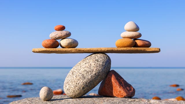 Mehrere Steine sind in Form einer ausgeglichenen Waage aufgeschichtet. Im Hintergrund sieht man das Meer und den blauen Himmel.