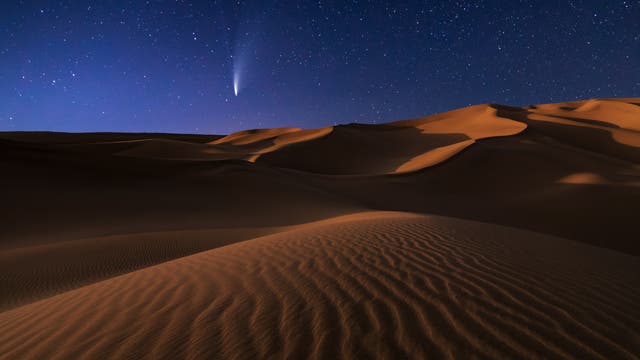 Eine nächtliche Wüstenlandschaft mit sanften Sanddünen im Vordergrund, beleuchtet vom Mondlicht. Am klaren Sternenhimmel ist ein heller Komet mit einem langen Schweif zu sehen, der von links oben nach rechts unten verläuft. Die Szene vermittelt eine ruhige und majestätische Atmosphäre.