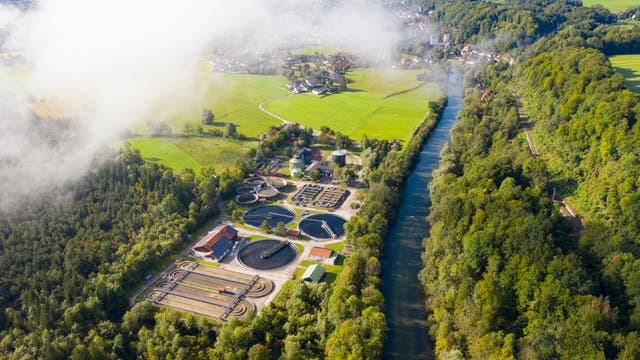 Luftaufnahme einer Kläranlage inmitten einer grünen Landschaft. Die Anlage besteht aus mehreren runden Becken und Gebäuden, die von Bäumen umgeben sind. Ein Fluss fließt rechts parallel zur Anlage, während im Hintergrund Felder und eine kleine Siedlung zu sehen sind. 