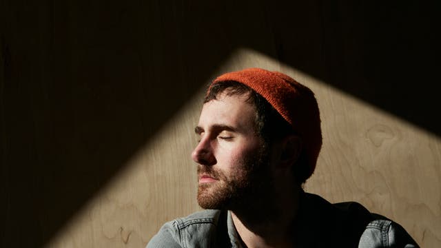 Ein Mann mit Jeanshemd und einer roten Strickmütze im Halbprofil genießt mit geschlossenen Augen das Sonnenlicht, das auf sein Gesicht scheint. 
