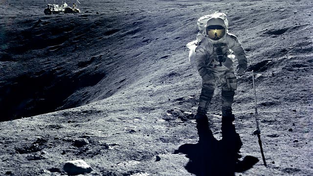 Ein Astronaut in einem Raumanzug steht auf der Mondoberfläche, umgeben von grauem, staubigem Terrain. Im Hintergrund ist ein Mondfahrzeug zu sehen. Der Astronaut hält einen langen Stab und wirft einen Schatten auf den Boden. Der Himmel ist schwarz, ohne sichtbare Sterne. Fußabdrücke sind im Vordergrund erkennbar.