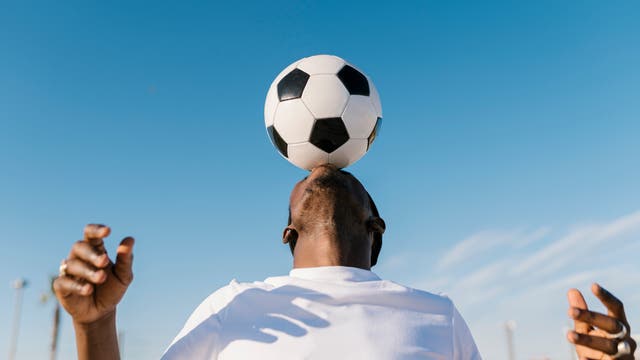 Eine Person balanciert einen Fußball auf der Stirn unter einem klaren blauen Himmel. Die Person trägt ein weißes T-Shirt und hebt die Hände leicht an, um das Gleichgewicht zu halten. Im Hintergrund sind verschwommene Strukturen und Lichtmasten zu sehen.