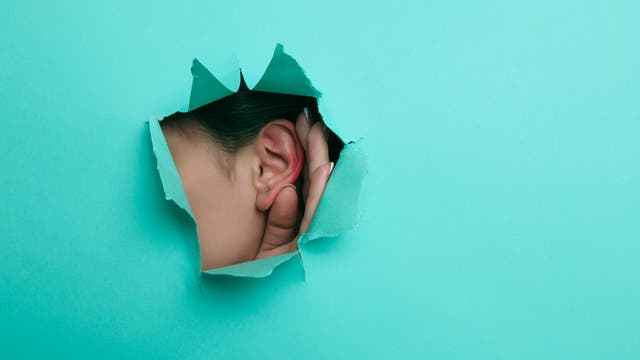 Eine Person hält ihre Hand an ihr Ohr und lauscht durch ein Loch in einer türkisfarbenen Papierwand. Das Bild symbolisiert das Konzept des Zuhörens oder Lauschens.