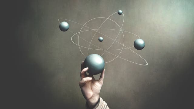 Eine Hand hält eine große, silberne Kugel, umgeben von kleineren Kugeln und weißen Linien, die an ein Atommodell erinnern. Der Hintergrund ist schlicht und dunkel, wodurch die Kugeln hervorgehoben werden. Die Szene vermittelt ein Gefühl von Wissenschaft und Technologie.