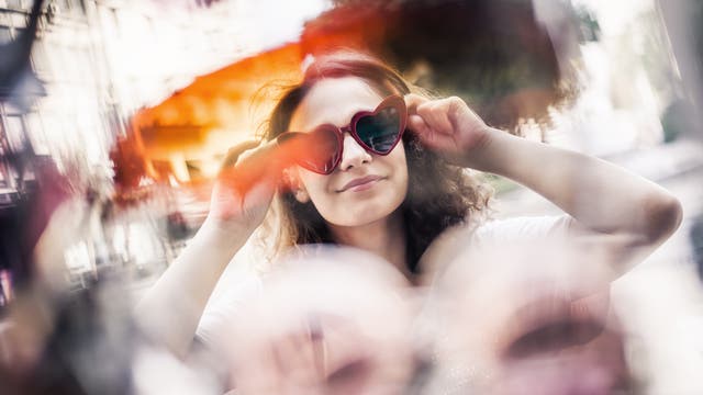 Eine Frau mit Locken trägt eine herzförmige Sonnenbrille und lächelt leicht. Die Umgebung ist verschwommen, was einen künstlerischen Effekt erzeugt. Die Farben sind warm und lebendig. Die Szene vermittelt ein Gefühl von Freude und Unbeschwertheit.