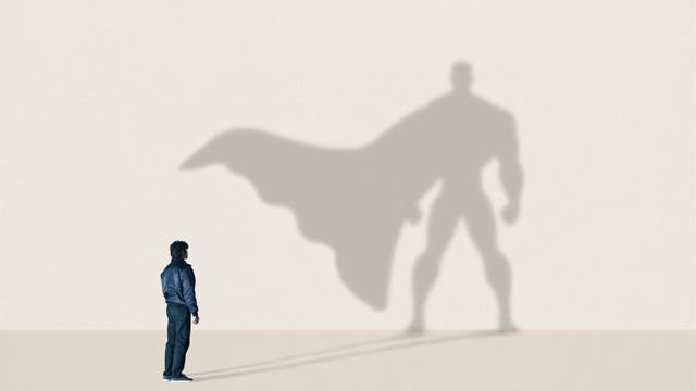 Eine Person steht vor einer Wand, auf der der eigene Schatten als großer Superheld mit Umhang zu sehen ist. Die Szene vermittelt das Wunschdenken von innerer Stärke und Potenzial. Der Hintergrund ist schlicht und hell, wodurch der Schatten deutlich hervorsticht.