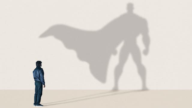 Eine Person steht vor einer Wand, auf der der eigene Schatten als großer Superheld mit Umhang zu sehen ist. Die Szene vermittelt das Wunschdenken von innerer Stärke und Potenzial. Der Hintergrund ist schlicht und hell, wodurch der Schatten deutlich hervorsticht.