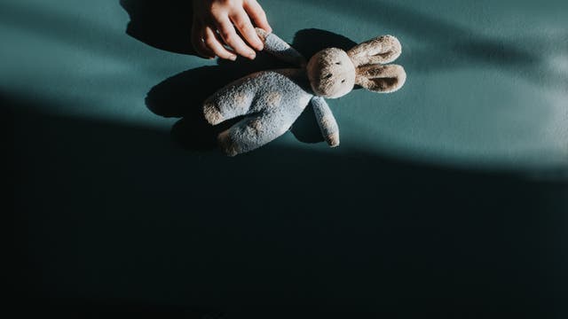 Eine Hand hält ein Plüschtier in Form eines Hasen auf einer dunkelgrünen Oberfläche. Das Bild ist von Schatten durchzogen, die eine ruhige und nachdenkliche Stimmung erzeugen. Der Hase ist aus weichem Stoff und hat lange Ohren.