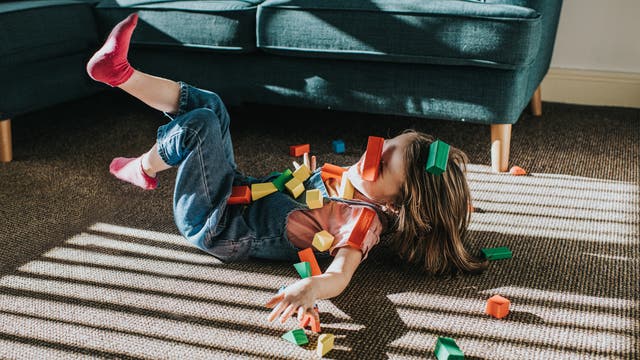 Ein Kind liegt lachend auf einem Teppichboden; bunte Holzbausteine liegen drumherum und auf dem Körper – es sieht so aus, als sei gerade ein Turm Bauklötzchen umgefallen und das Kind mit dazu. Das Kind trägt eine Jeans-Latzhose und rosarote Socken. Im Hintergrund steht ein blaues Sofa. Die Szene wirkt fröhlich und verspielt.