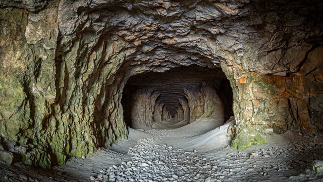 Ein langer, natürlicher Höhlentunnel mit unebenen, felsigen Wänden und einer steinigen, sandbedeckten Bodenfläche. Der Tunnel verjüngt sich in der Ferne und ist von weichem Licht erleuchtet, das die Textur der Felsen hervorhebt. Die Wände zeigen grünliche und bräunliche Farbtöne, die auf mineralische Ablagerungen hinweisen.