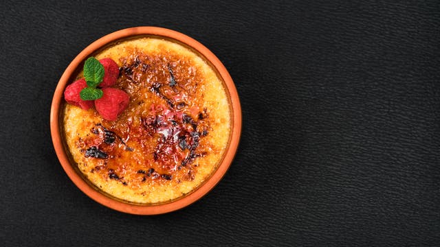 Eine Crème brulée in einer runden, braunen Schale auf schwarzem Hintergrund. Die karamellisierte Oberfläche ist goldbraun und knusprig. Oben links sind frische Himbeeren und ein Minzblatt zur Dekoration platziert.