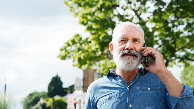 Ein älterer Mann steht im Freien und telefoniert mit einem Mobiltelefon. Er trägt ein blaues Hemd. Im Hintergrund sind Bäume und unscharfe Gebäude zu sehen, darunter eine britische Flagge. Der Himmel ist teilweise bewölkt.