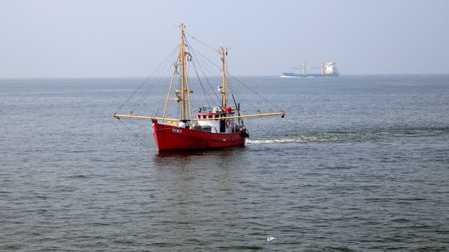 ein Foto zeigt einen roten Trawler mit ausgefahrenen Grundschleppnetzen in der Nordsee, im Hintergrund ist ein Frachter zu sehen