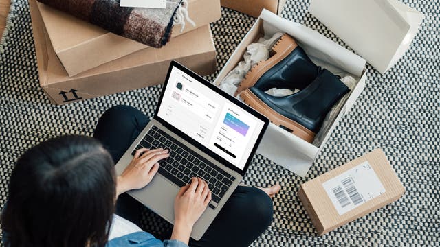 Eine Person sitzt auf einem Teppichboden und benutzt einen Laptop, um online einzukaufen. Auf dem Bildschirm ist eine Checkout-Seite zu sehen. Um die Person herum liegen geöffnete Pakete mit Schuhen und einem gefalteten Schal.