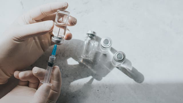 Eine Person mit medizinischen Handschuhen zieht Flüssigkeit aus einem Glasfläschchen in eine Spritze auf. Im Hintergrund sind weitere leere Fläschchen auf einer hellen Oberfläche zu sehen. Die Szene ist gut beleuchtet und vermittelt einen klinischen, sterilen Eindruck.