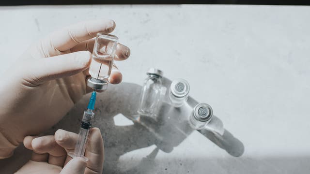 Eine Person mit medizinischen Handschuhen zieht Flüssigkeit aus einem Glasfläschchen in eine Spritze auf. Im Hintergrund sind weitere leere Fläschchen auf einer hellen Oberfläche zu sehen. Die Szene ist gut beleuchtet und vermittelt einen klinischen, sterilen Eindruck.