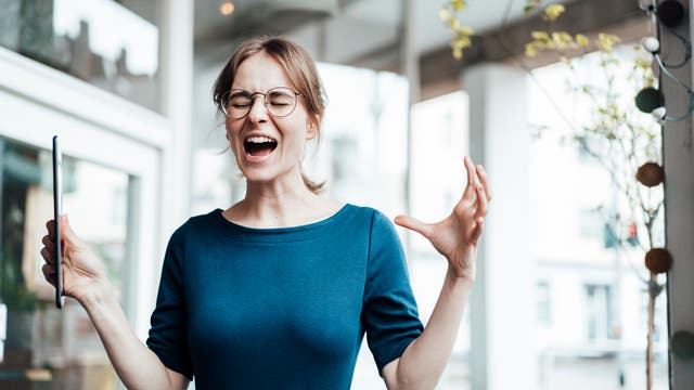 Eine junge Frau in einem blauen Oberteil steht in einem modernen Büro. Sie hält beide Arme leicht erhoben, in einer Hand ein Tablet. Ihre Mimik wirkt gequält und angestrengt: Sie hat die Augen zusammengekniffen und den Mund weit offen, als würde sie schreien.