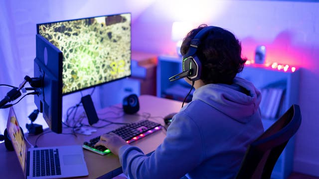 Eine Person sitzt an einem Schreibtisch und spielt ein Computerspiel. Sie trägt ein Headset und schaut auf einen großen Monitor, der ein Spiel mit einer detaillierten Karte zeigt. Auf dem Schreibtisch befinden sich eine beleuchtete Tastatur, eine Maus, ein Laptop und ein Mikrofon. Der Raum ist in blauem und violettem Licht beleuchtet, was eine Gaming-Atmosphäre schafft. Im Hintergrund sind Regale mit Büchern und Dekorationen zu sehen.