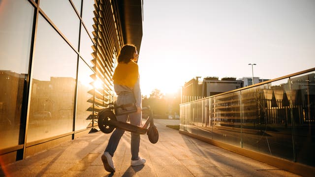 Eine Person geht bei Sonnenuntergang entlang eines modernen Gebäudes mit Glasfassade. Sie trägt einen zusammengeklappten Elektroroller und hat eine gelbe Jacke über die Schultern gelegt. Die Szene ist in warmes, goldenes Licht getaucht, das lange Schatten auf den Boden wirft. Im Hintergrund sind städtische Strukturen und ein klarer Himmel zu sehen.