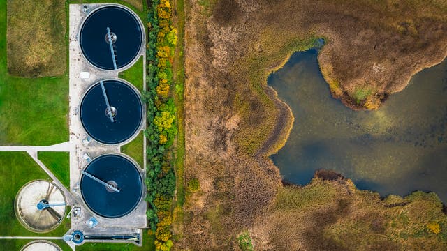 Luftaufnahme einer Kläranlage mit vier großen, runden Becken auf der linken Seite, umgeben von grünen Rasenflächen und Bäumen. Rechts davon erstreckt sich eine natürliche Landschaft mit einem Teich und Schilfgebieten. Die Anlage zeigt den Kontrast zwischen menschlicher Infrastruktur und Natur.