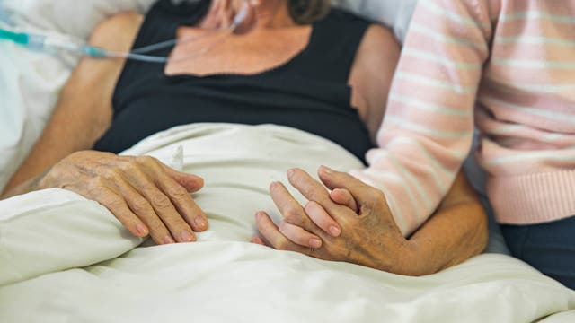 Eine ältere Frau liegt im Krankenhausbett, mit einer Sauerstoffnasenbrille verbunden. Ein Kind sitzt neben ihr und hält sanft ihre Hand.