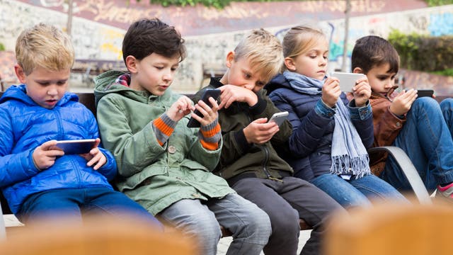 Fünf Kinder sitzen auf einer Bank im Freien und konzentrieren sich auf ihre Smartphones. Sie tragen Jacken. Im Hintergrund ist eine mit Graffiti bedeckte Wand zu sehen. Jedes Kind ist in sein Handy vertieft, was auf die Nutzung von Technologie in der Freizeit hinweist.