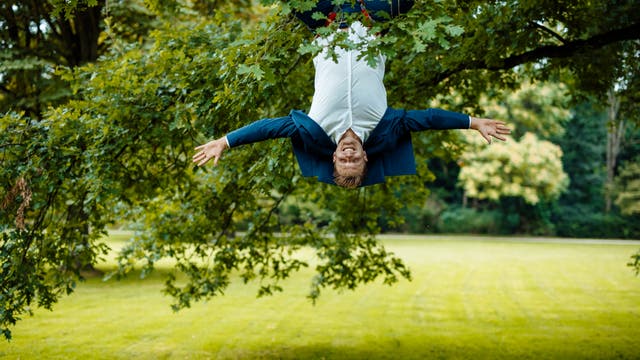 Ein Mann in weißem Hemd und einem blauen Anzug hängt kopfüber an einem Baum in einem Park. Er lächelt und breitet die Arme aus. Im Hintergrund sind grüne Wiesen und Bäume zu sehen.
