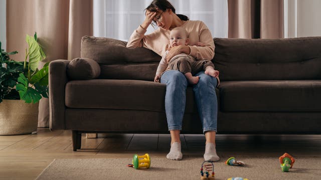 Eine Mutter sitzt auf einem Sofa und hält ihr Baby, sie sieht müde und gestresst aus.