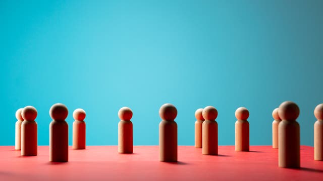 Holzfiguren stehen auf einer roten Oberfläche vor einem blauen Hintergrund. Die Figuren sind gleichmäßig verteilt und symbolisieren möglicherweise soziale Interaktion oder Gruppendynamik. Die Szene ist minimalistisch und kontrastreich gestaltet.