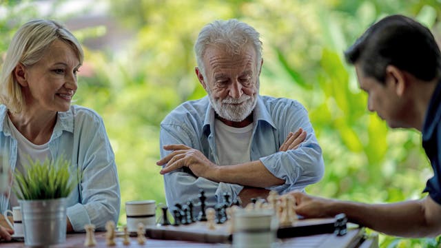Drei Personen sitzen an einem Tisch im Freien und spielen Schach. Eine ältere Frau und ein älterer Mann beobachten konzentriert das Spiel, während ein jüngerer Mann einen Zug macht. Im Vordergrund steht eine Topfpflanze auf dem Tisch. Der Hintergrund ist grün und verschwommen, was auf einen Garten oder Park hindeutet.