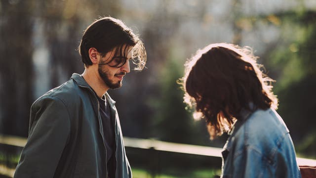 Zwei junge Menschen stehen in einem Park. Die Person links blickt nach unten, während die Person rechts leicht nach vorne geneigt ist. Beide tragen lässige Jacken. Die Szene ist von einem warmen Sonnenlicht durchflutet, das die Umgebung und die Personen sanft beleuchtet. Im Hintergrund sind unscharf Bäume und ein Geländer zu sehen.