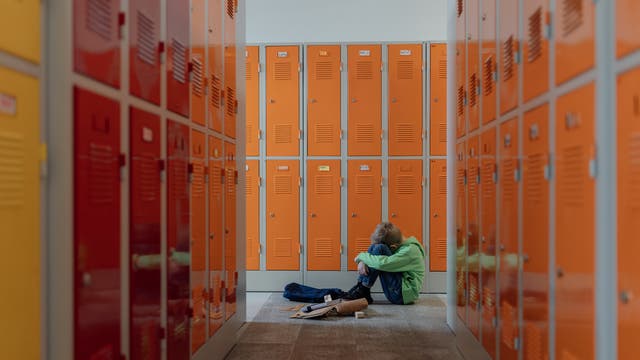 Ein Kind sitzt allein auf dem Boden eines Flurs mit orangefarbenen Schließfächern. Es hat die Knie angezogen und den Kopf darauf gelegt. Neben ihm liegen verstreute Bücher und eine Tasche. Die Szene vermittelt ein Gefühl von Einsamkeit und Nachdenklichkeit.