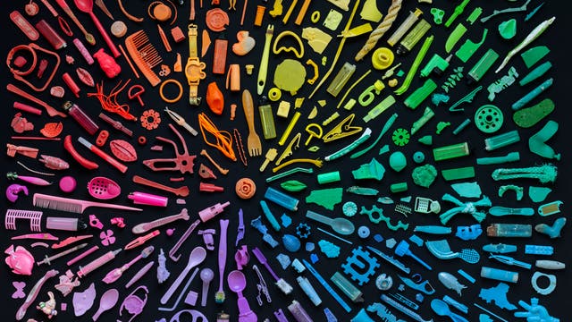 Eine Sammlung von Plastikgegenständen in verschiedenen Formen und Größen, die in einem Regenbogenmuster auf einem schwarzen Hintergrund angeordnet sind. Die Objekte sind in Farbtönen von Rot, Orange, Gelb, Grün, Blau bis Violett sortiert. Zu den Gegenständen gehören Kämme, Flaschenverschlüsse, Spielzeugteile und Besteck. Diese Anordnung zeigt die Vielfalt und Menge an Plastikmüll.