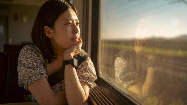 Asiatisch aussehende Frau mit schwarzen Haaren und einem geblühmten Sommerkleid sitzt in einem Zug und schaut aus dem Fenster auf die vorbeiziehende Landschaft, in der gerade die Sonne untergeht.