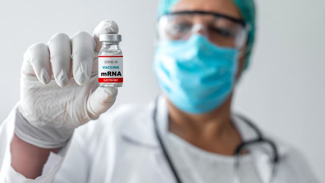 Eine Person in medizinischer Schutzkleidung, einschließlich Maske und Handschuhen, hält ein Fläschchen mit der Aufschrift „COVID-19 Vaccine mRNA“ in die Kamera. Der Hintergrund ist unscharf, der Fokus liegt auf dem Impfstofffläschchen.