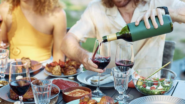 Eine Gruppe von Menschen sitzt an einem Tisch im Freien und genießt ein gemeinsames Essen. Ein Mann schenkt Rotwein in ein Glas ein. Auf dem Tisch stehen verschiedene Speisen wie gegrilltes Fleisch, Salat und Brot.