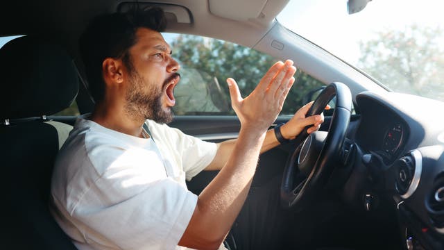 Ein Mann sitzt am Steuer eines Autos und zeigt eine frustrierte oder verärgerte Geste mit der rechten Hand erhoben. Er trägt ein weißes T-Shirt und hat dunkles Haar und einen Bart. Die Sonne scheint durch das Autofenster, und im Hintergrund sind verschwommene Bäume zu sehen.